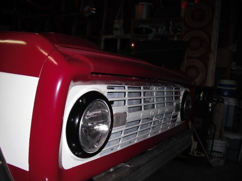 1968 Ford Bronco - Tight low-angle close-up of the front corner of a red early Bronco, showing the white-painted horizontal-bar grille with embossed FORD lett…