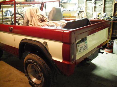 1968 Ford Bronco - Driver-side 3/4 rear view of a topless red-and-white early Bronco inside a workshop, showing the tailgate with a California plate reading 4…