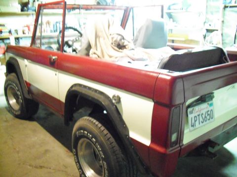 1968 Ford Bronco - Driver-side rear three-quarter view of a two-tone red-over-white early Ford Bronco in a garage, topless with a tan drop cloth thrown over t…