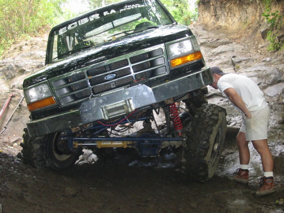 Dark green lifted Ford 4x4 with winch climbing a muddy rock ledge as a man spots