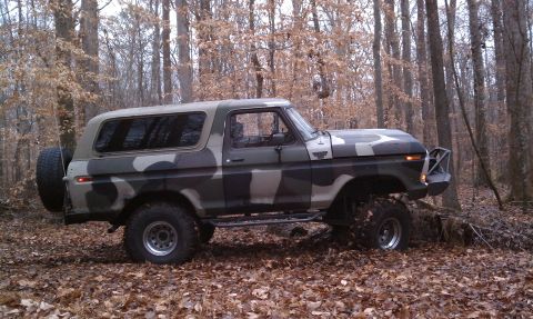 1979 Ford Bronco - A camouflaged Ford Bronco sits in a forest, its tires elevated on uneven terrain.