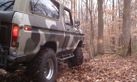 1979 Ford Bronco - This camouflaged Ford Bronco is navigating a wooded area with fallen leaves on the ground.