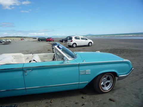 1964 Mercury Monterey - A turquoise convertible sits on a sandy beach with other vehicles in the background under a bright blue sky.