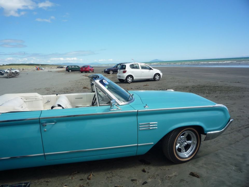Turquoise convertible with white interior on a beach, chrome trim, side vents, and aftermarket wheels