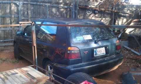 1991 Ford Festiva - This dark hatchback is parked in a yard with a wooden fence and some scrap metal structures.