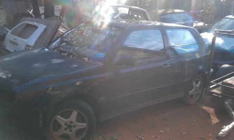 1991 Ford Festiva - A dark hatchback, likely a 1993 Volkswagen Golf, sits in a junkyard with other vehicles visible.