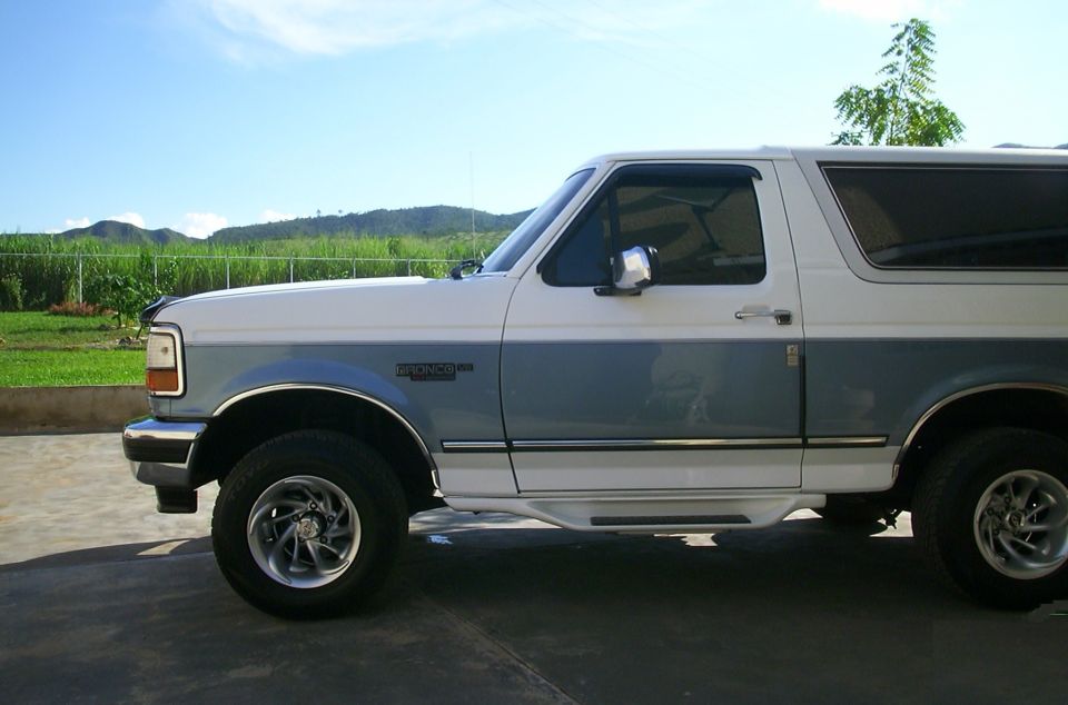 Side view of a white and blue 1993 Ford Bronco parked on pavement with a grassy hill and trees in the backgro…