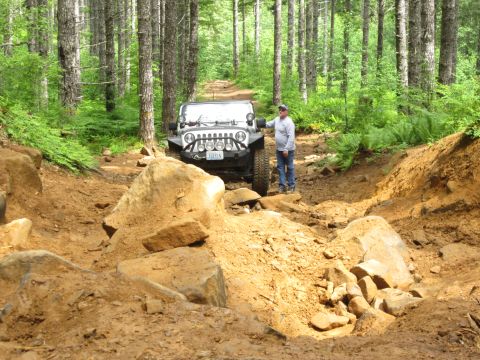 2008 Jeep Wrangler - A man stands beside a Jeep Wrangler navigating a challenging, rocky off-road trail through a forest.