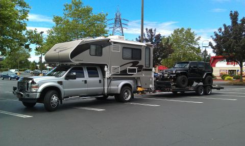 2008 Jeep Wrangler - A large truck carries a camper and tows a Jeep Wrangler on a trailer in a parking lot.
