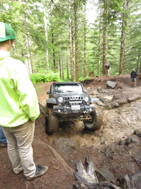 2008 Jeep Wrangler - A Jeep Wrangler tackles a challenging muddy obstacle on a wooded trail, with onlookers observing the off-road adventure.