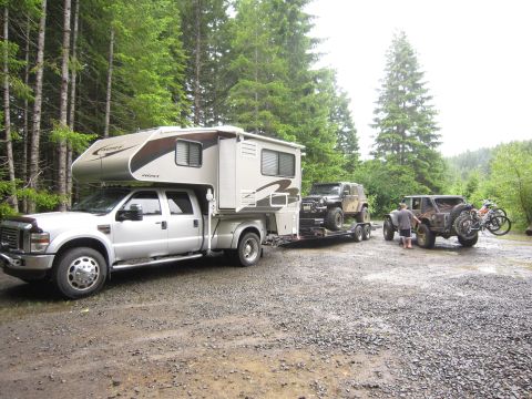 2008 Jeep Wrangler - A silver Ford truck tows a Jeep Wrangler on a trailer, with another Jeep and motorcycle parked nearby in a wooded area.