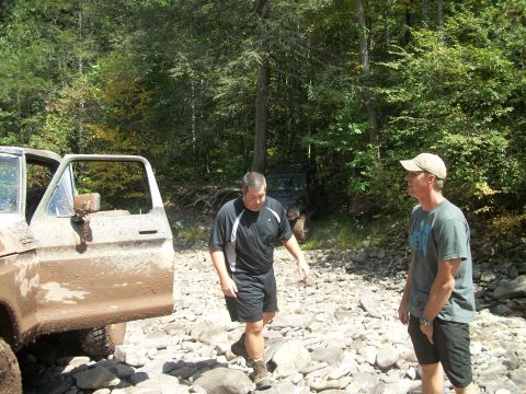 1979 Ford Bronco - Two men stand near a mud-covered 1977 Ford F-150 in a wooded, rocky area with another vehicle visible.