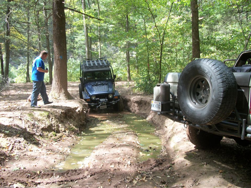 Two off-road vehicles, one blue Jeep and a rear view of another vehicle, drive through a muddy, wooded trail
