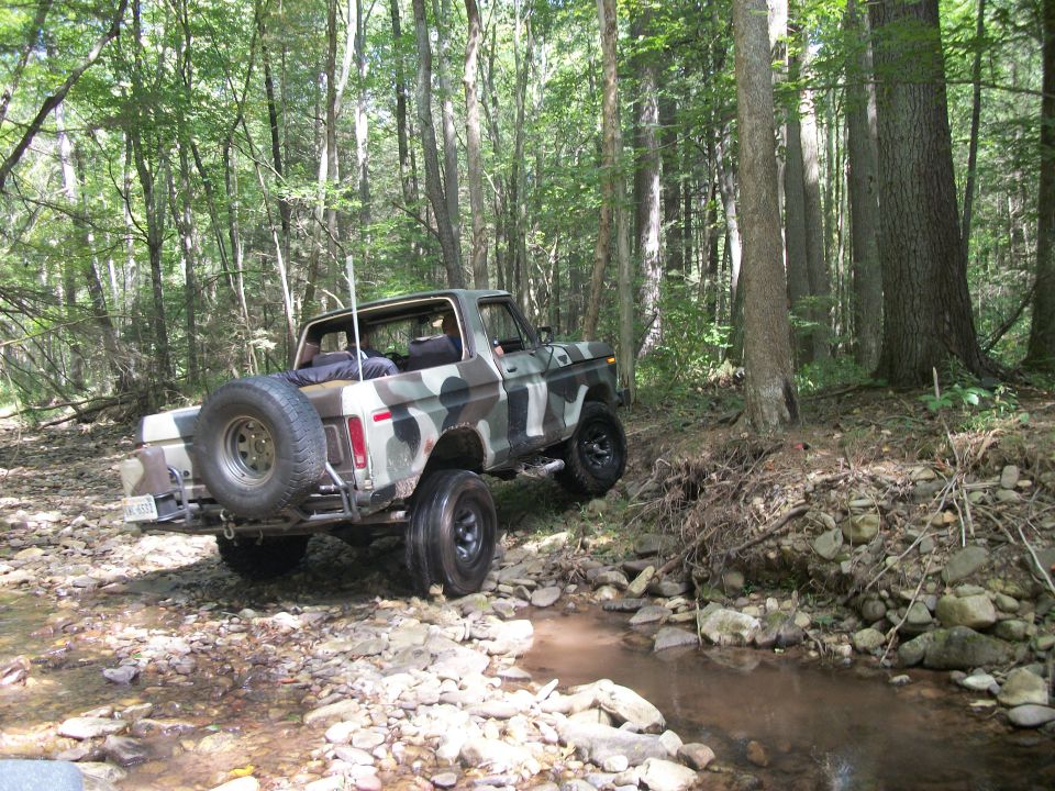 Camouflaged Ford Bronco driving through a rocky stream in a forest. Spare tire mounted on the rear