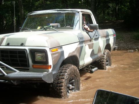 1979 Ford Bronco - This camouflage-painted Ford pickup truck is navigating through a shallow body of water in a wooded area.