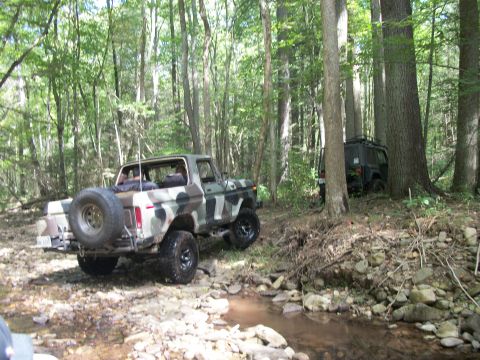 1979 Ford Bronco - A camouflaged Ford Bronco navigates a wooded stream, with another dark SUV visible behind it.