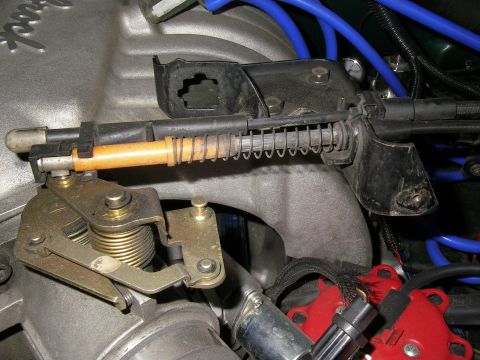 1990 Ford Bronco - Close-up of an engine intake area showing a copper throttle linkage rod, return spring, brackets and blue spark plug wires.