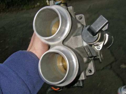 1990 Ford Bronco - Hand holding an aluminum dual-bore throttle body showing two throttle bores, linkage and sensor on pavement.
