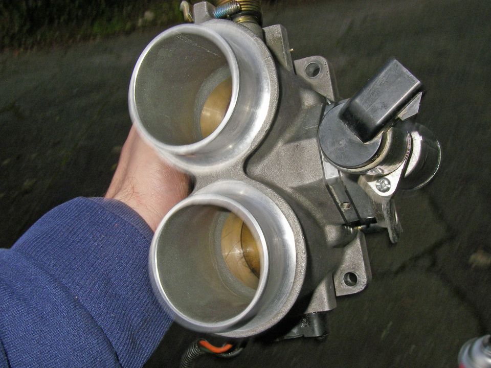Hand holding a dual-bore aluminum throttle body with throttle linkage
