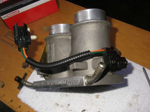 1990 Ford Bronco - A BBK Power-Plus twin 65mm aluminum throttle body sitting on a shop towel over a wood workbench, with a black wiring harness (orange/green…