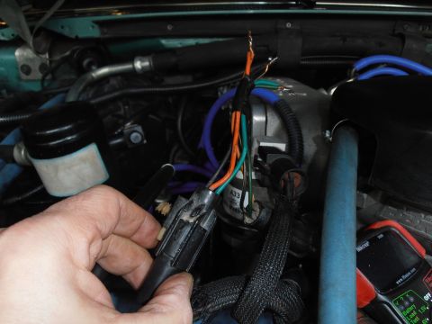 1990 Ford Bronco - Close-up of a hand holding a disconnected wiring harness in an engine bay, revealing exposed wires and a visible part number.