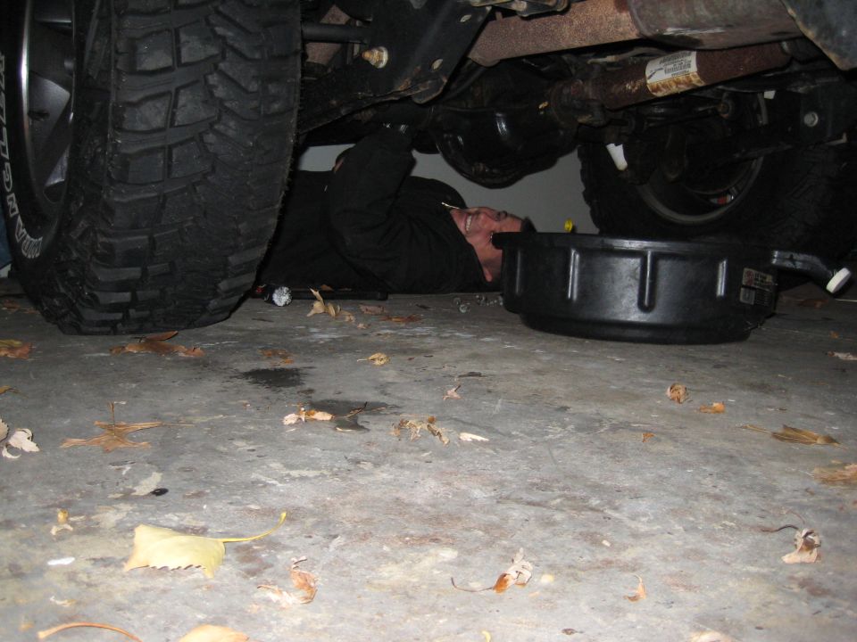 Person in black clothing lying on concrete floor under a vehicle, with a black oil drain pan nearby. Large tire visible on left