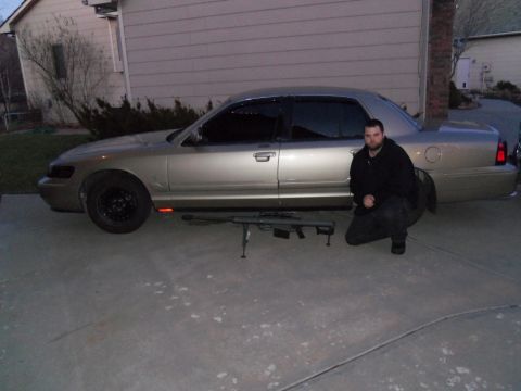 2000 Mercury Grand Marquis - A man kneels beside a gold 2000 Mercury Grand Marquis sedan, with a rifle placed in front of the vehicle.