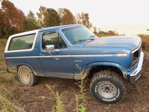 1985 Ford Bronco - This blue Ford Bronco features a white removable top and off-road tires, parked in a field during autumn.