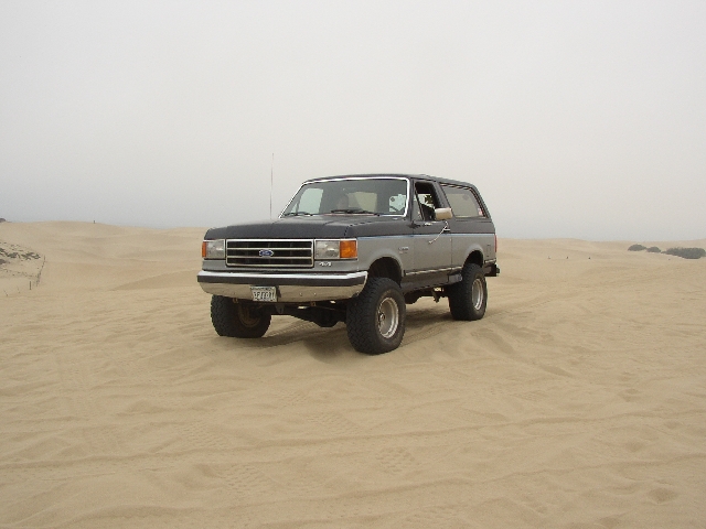 Gray and silver lifted Ford Bronco SUV parked on sand dunes, 3/4 front view.