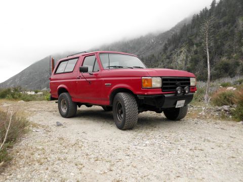 1988 Ford Bronco - Passenger-side three-quarter front view of a red Ford Bronco with a hardtop and roof rails parked on a gravel pullout, sporting a black tub…