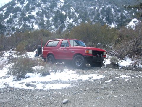 1988 Ford Bronco - A red 1988 Ford Bronco is parked on a dirt road with snow on the ground and trees in the background.