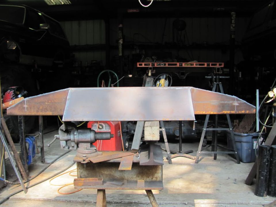 Metal bumper section clamped in a vise, with fabrication tools and a vehicle in the background