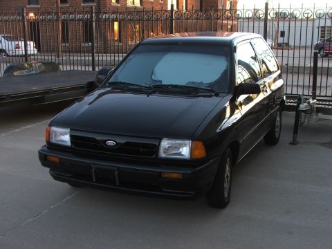 1991 Ford Festiva - This black hatchback is parked on a concrete lot with a metal fence in the background.