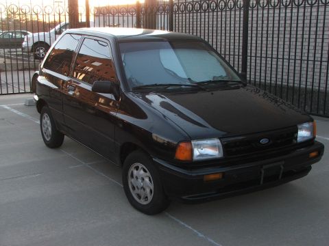 1991 Ford Festiva - A black 1991 Ford Festiva hatchback is parked in front of a metal fence.