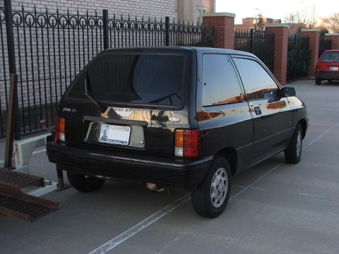 1991 Ford Festiva - This black 1991 Ford Festiva is parked outdoors next to a fence, with another red car visible in the background.