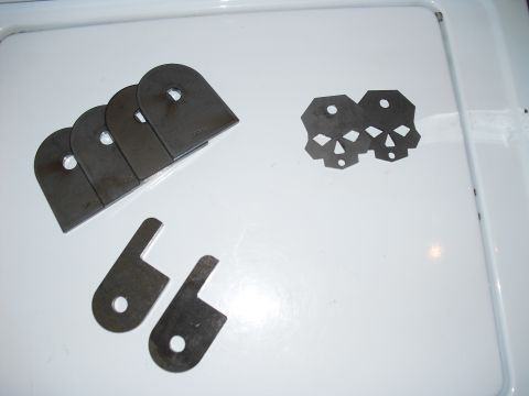 1986 Ford Bronco - Various metal fabrication parts including tab mounts, brackets, and skull cutouts are laid out on a white surface.