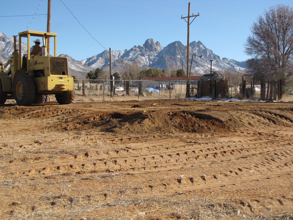 Yellow front loader with a person in the cab, seen from the side, operating on a dirt construction site with mountains in the background