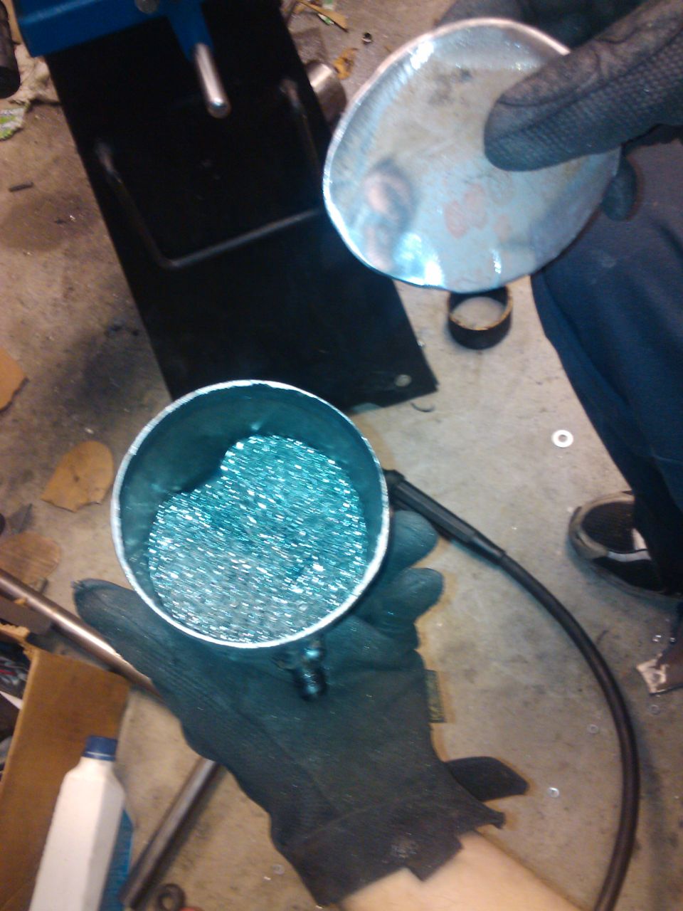 Gloved hands hold a metal cylinder filled with blue-tinted metal shavings and a detached metal cap