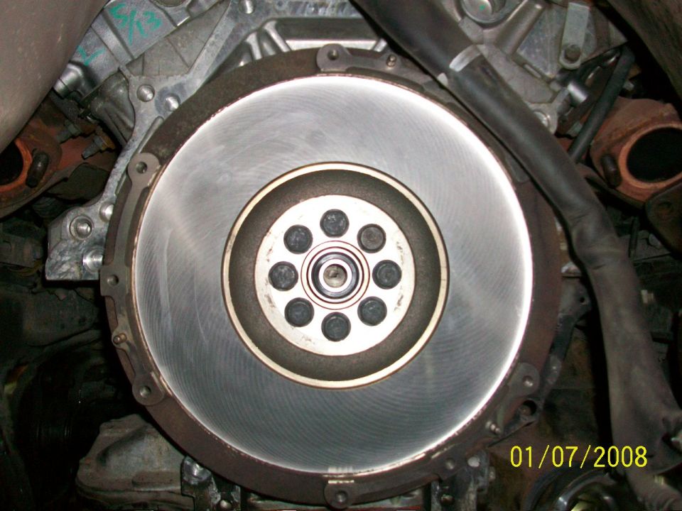 Close-up of a car's engine flywheel with bolts and a bearing visible. There's a date stamp in the corner