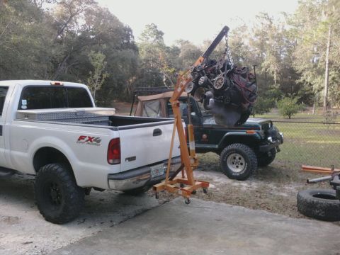 1995 Ford Bronco - An engine hoist is used to lift an engine out of a green Jeep, with a white pickup truck parked nearby.