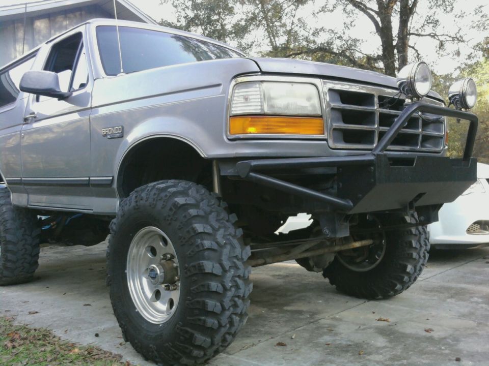 Low front view of lifted gray Ford Bronco with black tube bumper, driving lights and mud tires