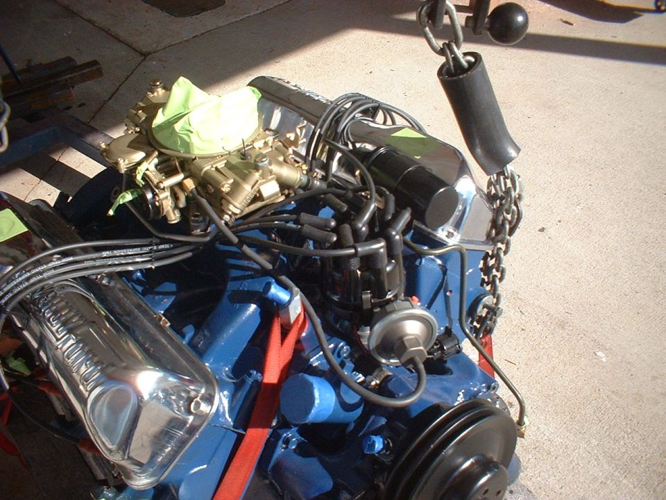 Engine block painted blue, with chrome valve covers, carburetor, and distributor. Being lifted by a chain hoist