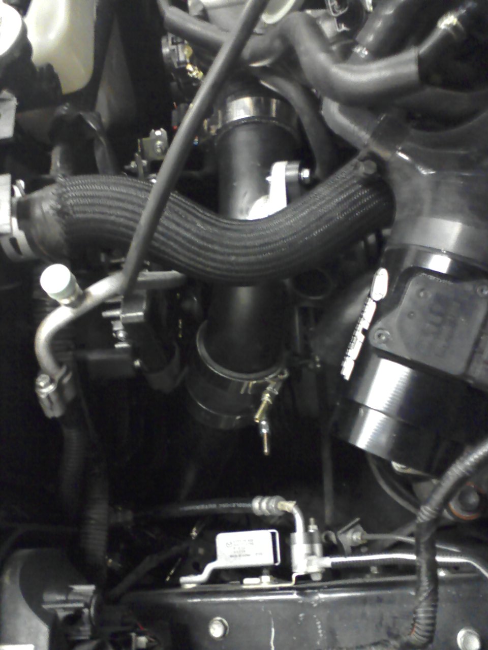 Close-up of engine bay with black hoses, pipes, and a silver component. Text on a white label is visible