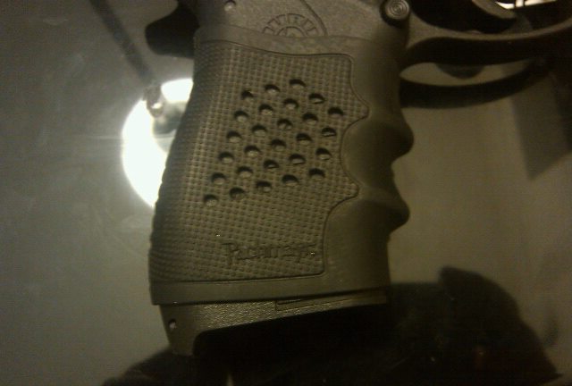 Close-up of a black pistol grip with textured surface and circular indentations. Text is visible on the grip