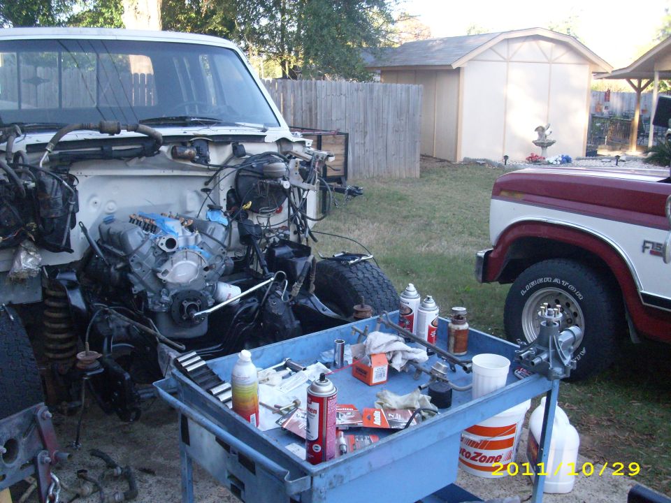 1990 Ford F-150 engine bay with engine removed, parts on a cart, and another F-series truck in background