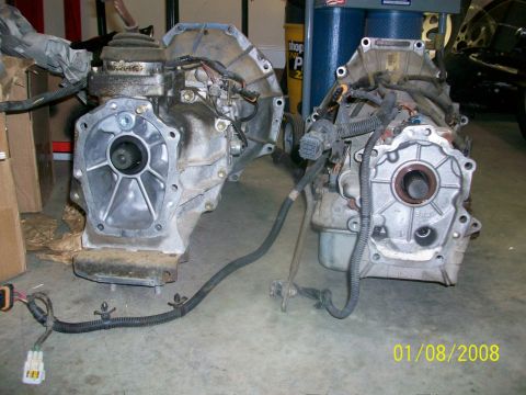 2000 Isuzu Vehicross - Two vehicle transmissions, likely for a 4x4, are displayed on a garage floor with visible wiring harnesses.