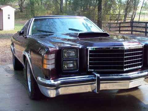 1979 Ford Ranchero - Deep maroon Ford coupe-utility shot from the front three-quarter angle on a wet concrete pad, with woods and a board fence behind. It has s…