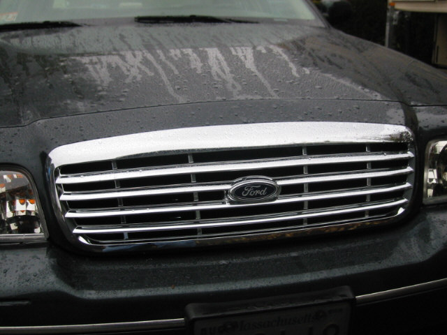 Front grille and headlights of a dark-colored Ford Crown Victoria. The grille has horizontal chrome bars and a Ford logo in the center. The hood has water droplets on it