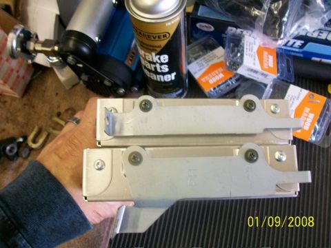 2000 Isuzu Vehicross - A person holds two metal brackets, with various automotive parts and hardware visible in the background.