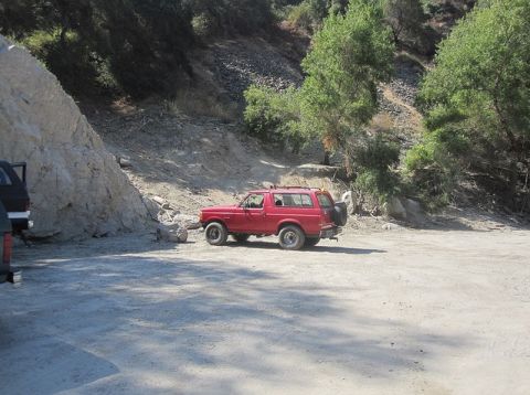 1988 Ford Bronco - A red SUV is parked on a dusty, unpaved road with a rocky incline and trees in the background.
